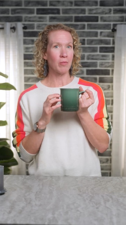Heather holding a green mug and looking straight into the camera on a brick-wall studio set